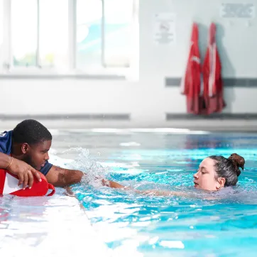Lifeguard lies down on the edge of an indoor pool and reaches out to assist a swimmer